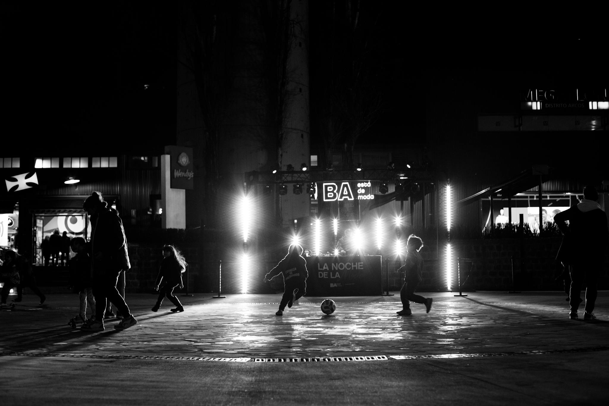 Black and white image of children playing soccer under city lights at night.