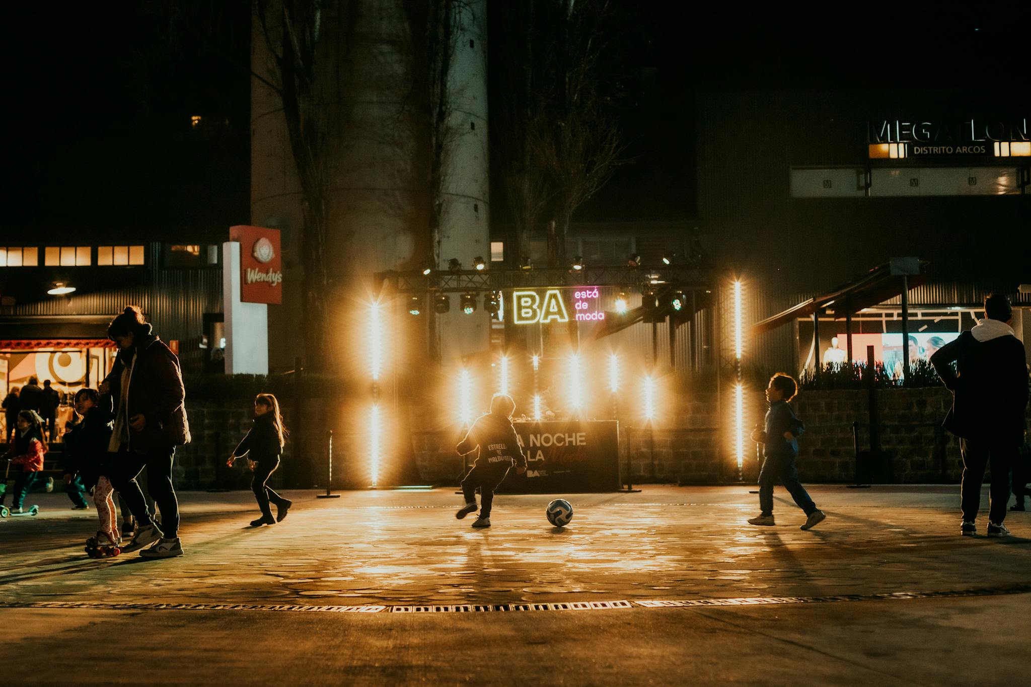Children enjoying a night soccer game at an outdoor urban event with festive lighting.